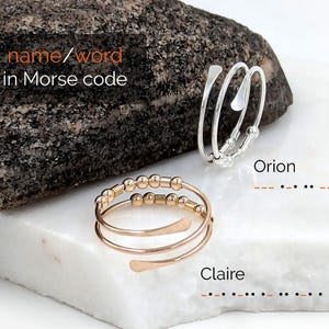 Könnte beinhalten: Zwei verstellbare Ringe, einer silberfarben und einer goldfarben, auf einer weißen Oberfläche. Der goldfarbene Ring hat kleine Perlen und der silberfarbene Ring ein minimalistisches Design. Der Text lautet "Name/Wort in Morsecode" und die Namen "Orion" und "Claire" mit ihrem Morsecode.