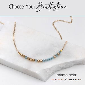 May include: A gold chain necklace featuring gold and light blue beads. The necklace is presented on a white marble surface. The text "Choose Your Birthstone" is at the top, and "mama bear" is at the bottom.