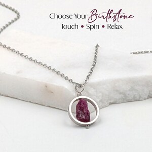 Pode incluir: Um colar de prata com um pingente com uma pedra preciosa vermelha escura bruta dentro de uma moldura circular. O texto "Choose Your Birthstone" é exibido acima do colar, com as palavras "Touch • Spin • Relax" abaixo.