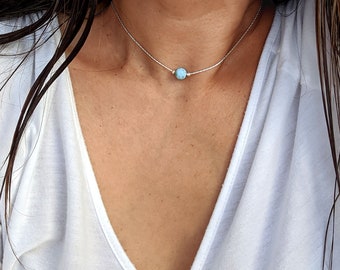May include: A delicate silver necklace featuring a small, round, light blue bead as a focal point. The necklace is worn against a white top, creating a simple and elegant look. The chain is thin and subtle.