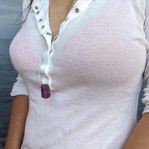 May include: A white long-sleeved shirt with a button-up front and a silver chain necklace with a pink stone pendant.