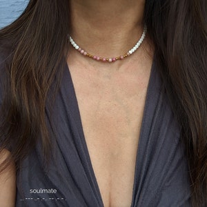 May include: A delicate choker necklace with white lava beads and pink gemstone beads. The necklace is worn by a person with long brown hair.