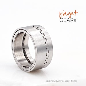 Op de afbeelding: Een zilveren fidgetring met een tandwielontwerp. De ring is gemaakt van metaal en heeft een geborstelde afwerking. "fidget GEARS" staat in oranje tekst boven de ring.