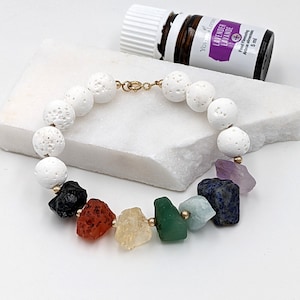 Może przedstawiać: Bransoletka z białych kamieni lawowych z pozłacanym zapięciem i siedmioma kolorowymi kamieniami szlachetnymi. Bransoletka leży na białym tle z butelką olejku eterycznego z lawendy.