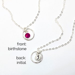 Könnte beinhalten: Eine silberne Kettenkette mit einem umkehrbaren Anhänger. Auf der einen Seite befindet sich ein kleiner roter Edelstein in einem Kreis, auf der anderen Seite ein silberner Kreis mit dem Buchstaben "J" in der Mitte. Die Kette ist mit "Reversible Kette" und "vorne: Geburtsstein, hinten: Initial" beschriftet.