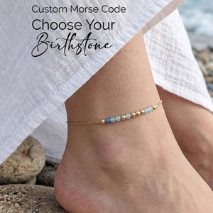 Puede incluir: Una delicada tobillera de cadena dorada con una serie de pequeñas cuentas redondas de color azul claro y dorado dispuestas en código Morse. El texto Custom Morse Code Choose Your Birthstone es visible en la esquina superior izquierda.