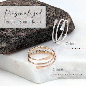 Puede incluir: Dos anillos en espiral, uno plateado y otro dorado, se exhiben sobre una superficie blanca. El anillo plateado tiene un diseño minimalista con pequeñas cuentas. El anillo dorado presenta una sección con cuentas y un diseño curvo. El texto "Personalized Touch Spin Relax" es visible.