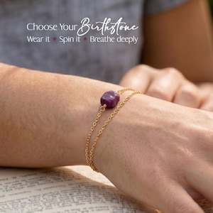 May include: A gold-colored chain bracelet with a large, rounded, purple gemstone. The bracelet is worn on a wrist, and the image includes the text "Choose Your Birthstone, Wear it, Spin it, Breathe deeply."