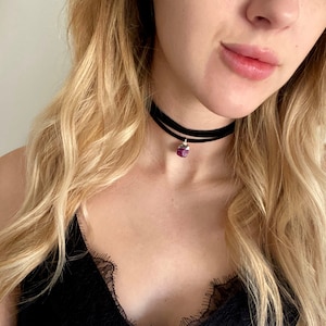 May include: A black choker necklace with a small, dark purple gemstone pendant. The necklace is worn by a person with long, blonde hair.
