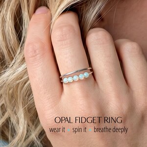 May include: A silver opal fidget ring with a double band design. The ring features five small, round opals set in a row. The text "OPAL FIDGET RING" is displayed below the ring, along with the phrases "wear it", "spin it", and "breathe deeply".