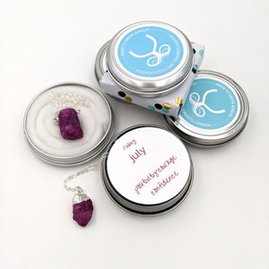 May include: A silver necklace with a raw ruby pendant in a small silver tin. The tin has a label that says "ruby july" and "positivity courage confidence".