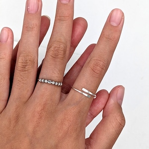 Two silver rings on a hand. One ring is a thin band with a series of small beads spelling an initial in Morse code. The other ring shows hammered paddle ends the bypass one another.