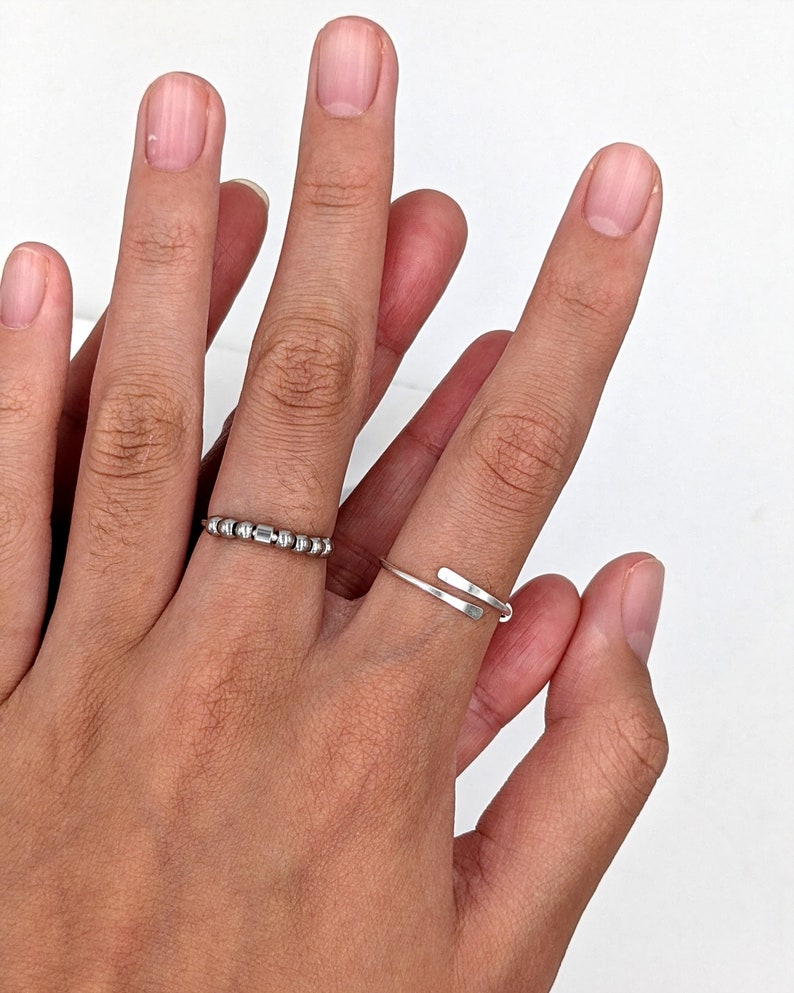Two silver rings on a hand. One ring is a thin band with a series of small beads spelling an initial in Morse code. The other ring shows hammered paddle ends the bypass one another.