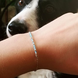 May include: Delicate bracelet featuring small, light blue beads and silver-colored accents. The bracelet is strung on a fine chain. The background includes a black and white dog.
