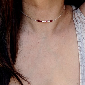 Könnte beinhalten: Eine zarte silberne Kettenkette mit kleinen roten und weißen Perlen. Die Kette ist im Choker-Stil gehalten und liegt eng am Hals an.