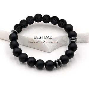 May include: A black beaded bracelet with the text "BEST DAD". The bracelet features a mix of matte black and textured black beads, along with silver-toned metal accents. The bracelet is displayed on a white surface.