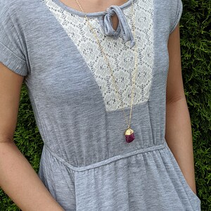 May include: A gray dress with a white lace overlay and a gold chain necklace with a dark purple pendant. The dress has a scoop neck and short sleeves.