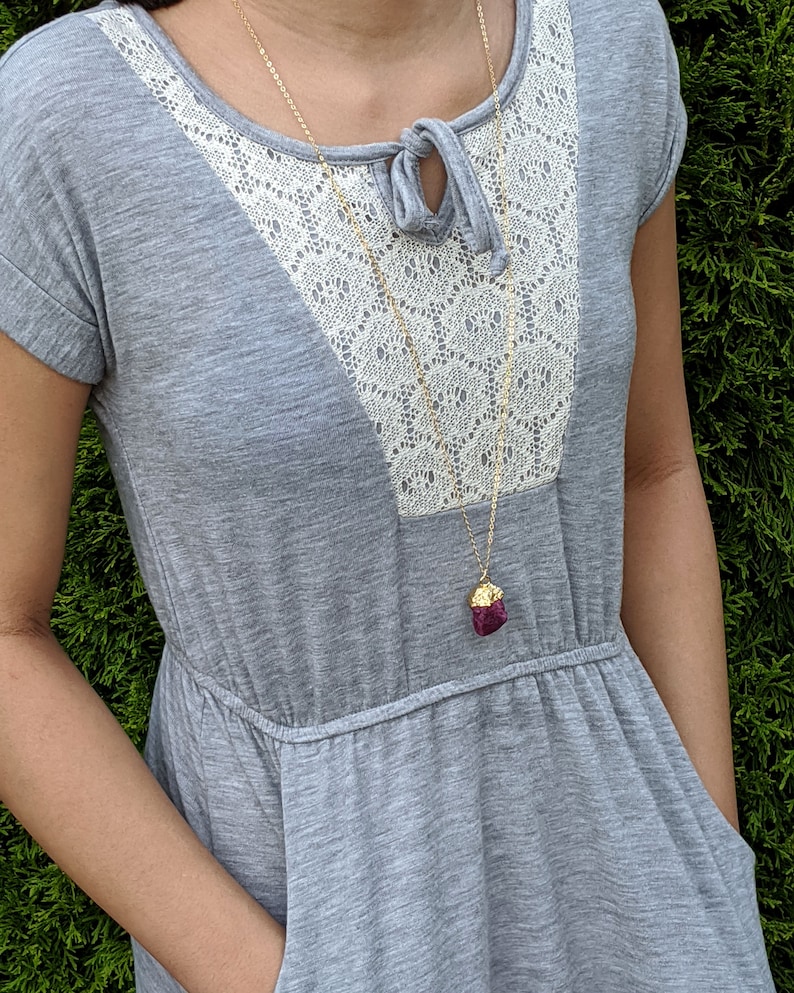 May include: A gray dress with a white lace overlay and a gold chain necklace with a dark purple pendant. The dress has a scoop neck and short sleeves.