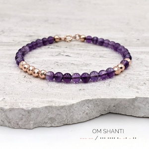 Op de afbeelding: Een delicate armband met amethist kralen en roségouden accenten. De armband bestaat uit kleine, ronde, dieppaarse amethist kralen afgewisseld met facet geslepen roségouden kralen. De sluiting is ook roségoud. De tekst "OM SHANTI" staat onderaan.