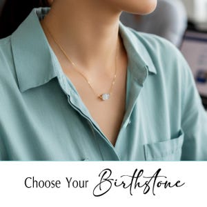 Op de afbeelding: Een delicate gouden ketting met een ronde, lichtgekleurde edelsteen hanger. De ketting wordt gedragen over een lichtblauw overhemd. De tekst "Choose Your Birthstone" staat onderaan de afbeelding.