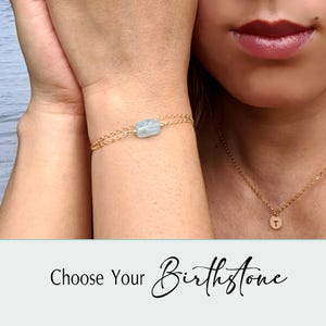 Puede incluir: Una pulsera dorada con una piedra preciosa azul claro y una fina cadena dorada. También se ve un collar dorado con un pequeño colgante redondo. El texto "Choose Your Birthstone" está en la parte inferior de la imagen.