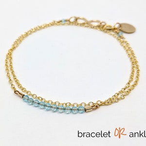 Könnte beinhalten: Ein zartes Armband aus goldener Kette mit einer Reihe hellblauer Perlen. Das Armband ist mit einer zweiten goldenen Kette übereinander gelegt. Der Text "Armband ODER Fußkettchen" ist unter dem Bild gedruckt.