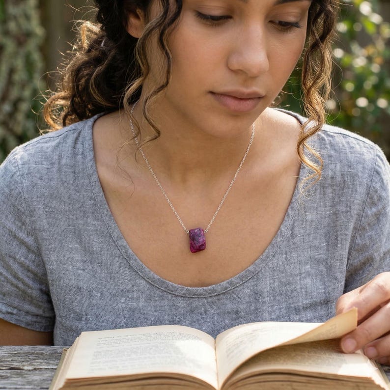 Close-up of a woman wearing a large raw ruby necklace on a silver chain, featuring a natural deep red gemstone pendant with an organic shape, styled casually while reading outdoors.