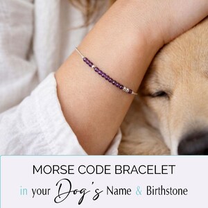 May include: A silver Morse code bracelet with purple beads and silver accents. The bracelet is worn on a person's arm, with a dog in the background. The text on the image reads "Morse Code Bracelet in your Dog's Name & Birthstone."