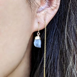 Può includere: Un paio di orecchini a catena dorata con un ciondolo di cristallo blu chiaro appeso a una catena. Gli orecchini sono mostrati nell'orecchio di una persona. Il testo "Style A" è visibile nella parte inferiore dell'immagine.