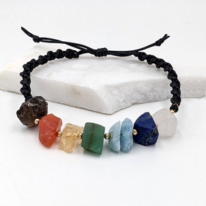 Könnte beinhalten: Ein Armband aus schwarzem Kordel mit sieben natürlichen Steinperlen in verschiedenen Farben, darunter klar, braun, orange, gelb, grün, blau und weiß.