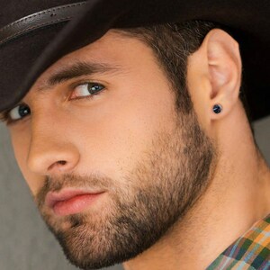 May include: A close-up of a man's face with a short beard. He is wearing a brown cowboy hat and a plaid shirt. He has a small, round, dark blue earring in his right ear.
