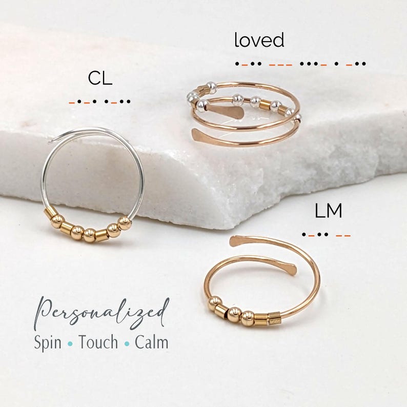 Personalized Morse code fidget rings displayed on a marble block, featuring silver and gold bead designs that spell words, names, and initials like “loved” and “LM,” with adjustable open bands made for spinning and calming stress relief.