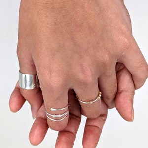 May include: Two hands wearing silver rings. One hand has a thick silver band on the thumb and a thin silver ring with three small beads on the middle finger. The other hand has a thin silver ring with gold beads on the ring finger.