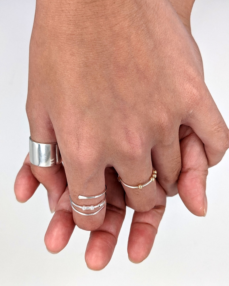 May include: Two hands wearing silver rings. One hand has a thick silver band on the thumb and a thin silver ring with three small beads on the middle finger. The other hand has a thin silver ring with gold beads on the ring finger.