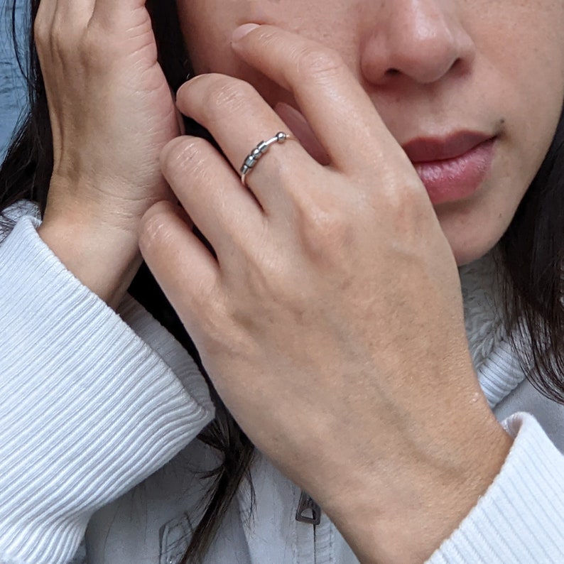 A silver ring with a row of small, round beads spelling an initial in Morse code. The ring is worn on the index finger of a person's hand.