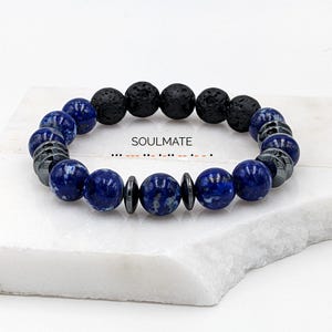 Pode incluir: Uma pulseira de contas com lápis-lazúli azul escuro, rocha vulcânica preta e contas de hematita cinza escuro. A palavra "SOULMATE" está impressa na superfície. Um acessório elegante.
