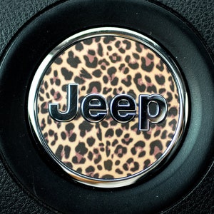 May include: A circular Jeep vehicle steering wheel emblem featuring a silver metallic Jeep logo centered over a tan and black leopard print background, all encased within a smooth silver ring set against a textured black steering wheel surface.