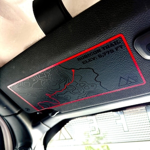 May include: A rectangular black decal with a red border applied to a vehicle sun visor. The decal features a topographic map design with a red trail line, the text "RUBICON TRAIL" and "ELEV: 6,775 FT" printed in black, and a mountain icon.