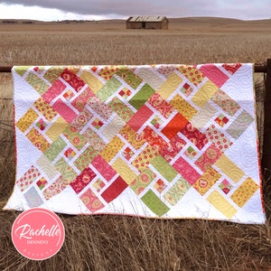 Könnte beinhalten: Ein farbenfroher Quilt mit einem geometrischen Muster aus Quadraten und Rauten in den Farben Rosa, Rot, Gelb, Grün und Weiß. Der Quilt hängt an einem Zaun vor einem Feld mit einem kleinen Gebäude in der Ferne. Der Quilt ist mit "Crossroads" und "Rachelle Dennery Designs" beschriftet, mit dem Text "Fat Eighths or Fat Quarter Friendly".