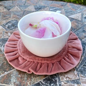 Op de afbeelding: Een witte theekop gevuld met roze en witte bloemblaadjes staat op een stoffig roze, gerimpelde onderzetter. De onderzetter staat op een mozaïek tafel, met witte en roze pioenrozen op de achtergrond.