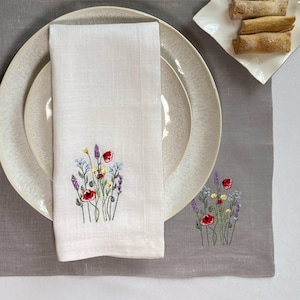 Può includere: Una tavola apparecchiata con un sottopiatto grigio, piatti bianchi e un tovagliolo di lino bianco ricamato con fiori di campo colorati. Posate d'argento, candele e un piattino con cibo sono anch'essi presenti.