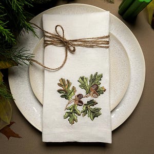 Può includere: Un tovagliolo di lino bianco con un ricamo a foglie di quercia e ghiande, legato con spago, è appoggiato su un piatto bianco. Il ricamo è verde, marrone e dorato. L'ambientazione suggerisce un tema autunnale.