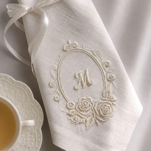 Puede incluir: Una servilleta de lino color blanco roto plegada que presenta un marco ovalado bordado con la letra M en el centro, rodeada de motivos florales de rosas. La servilleta est&aacute; atada con una fina cinta de sat&eacute;n blanco junto a una taza.
