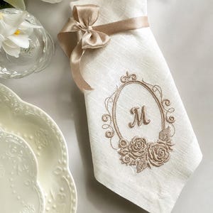Puede incluir: Una servilleta rectangular blanca con una cinta de sat&eacute;n beige atada en un lazo en la parte superior. El centro de la servilleta muestra un marco ovalado beige bordado con la letra M, acentuado con motivos florales de rosas en la parte inferior.