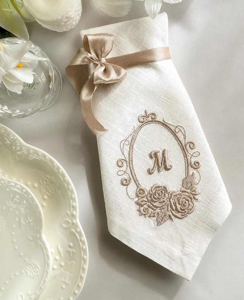 Puede incluir: Una servilleta rectangular blanca con una cinta de sat&eacute;n beige atada en un lazo en la parte superior. El centro de la servilleta muestra un marco ovalado beige bordado con la letra M, acentuado con motivos florales de rosas en la parte inferior.