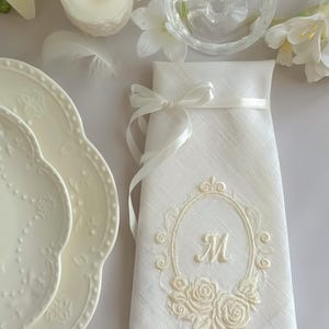 Puede incluir: Una servilleta de lino blanco con un marco ovalado bordado que presenta una letra M central y motivos florales de rosas en la base. La servilleta est&aacute; doblada verticalmente y atada con una fina cinta de sat&eacute;n blanco, creando una presentaci&oacute;n decorativa.