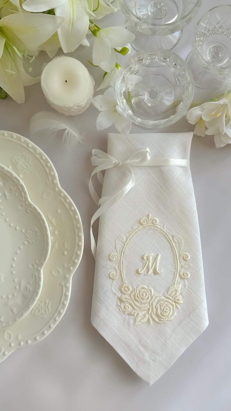 Puede incluir: Una servilleta de lino blanco con un marco ovalado bordado que presenta una letra M central y motivos florales de rosas en la base. La servilleta est&aacute; doblada verticalmente y atada con una fina cinta de sat&eacute;n blanco, creando una presentaci&oacute;n decorativa.
