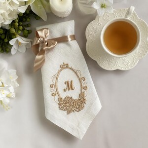 Puede incluir: Una servilleta de lino blanco con un marco ovalado bordado en hilo dorado, detalles florales y la letra M en el centro. La servilleta est&aacute; atada con una cinta de sat&eacute;n dorada y colocada junto a una taza de t&eacute; blanca y flores blancas.