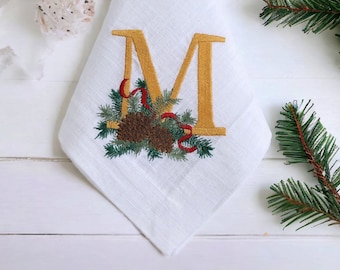 May include: A white linen napkin with an embroidered gold letter "M" surrounded by pine branches, pine cones, and red ribbon.