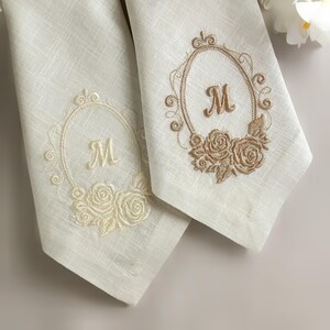Puede incluir: Dos servilletas de lino color blanco roto con monogramas bordados. Cada servilleta muestra la letra M centrada en un marco ovalado, decorado con motivos de rosas en la base. La servilleta izquierda tiene bordado crema y la derecha es beige.